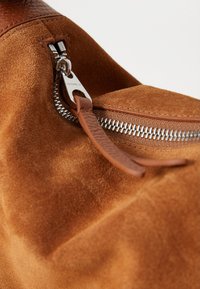 Coin du sac en suède marron avec une fermeture éclair en métal argenté. La tirette de la fermeture éclair est dotée d'une sangle en cuir et d'une plaque logo, mettant en valeur des matériaux texturés.