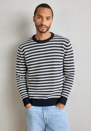 Hombre con un suéter de punto a rayas blancas y negras y jeans azules claros, con las manos en los bolsillos frente a un fondo gris claro.