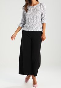 Chemise blanche à pois noirs, encolure froncée et manches trois-quarts, associée à un pantalon noir large taille haute et des talons roses.