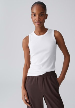 Vrouw met een donkere huid en haar naar achteren gebonden, draagt een witte mouwloze top en bruine broek met elastische taille, staat met één hand in de zak.