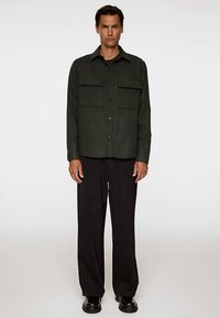 Mörkgrön skjorta med knappar och två framfickor, med krage och ett texturerat tyg. Matchad med svarta vidbyxor och skor.