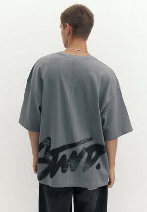 Homme portant un t-shirt gris oversized avec une grande impression noire style graffiti sur le bas du dos, associé à un pantalon sombre, dos à la caméra.