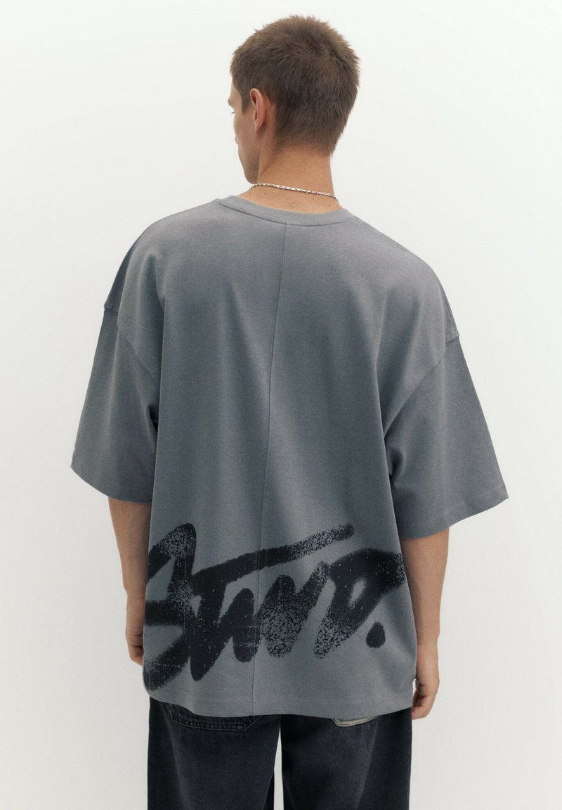 Homme portant un t-shirt gris oversized avec une grande impression noire style graffiti sur le bas du dos, associé à un pantalon sombre, dos à la caméra.