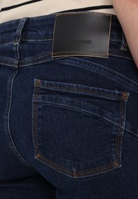 Poche arrière d’un jean en denim bleu foncé avec surpiqûres contrastantes orange et une étiquette de marque en cuir noir indiquant SPORTMAX à la taille.