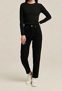 Personne portant un haut noir à manches longues, un pantalon noir taille haute et des baskets blanches, debout devant un fond neutre.