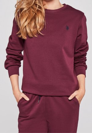 Kvinde iført en vinrød sweatshirt og matchende bukser med hænderne i lommerne, hår faldende over den ene skulder, ensfarvet baggrund.