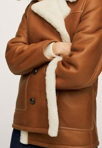 Persona che indossa una giacca di pelle marrone con rivestimento in shearling bianco, braccia incrociate e pantaloni scuri visibili nella parte inferiore.