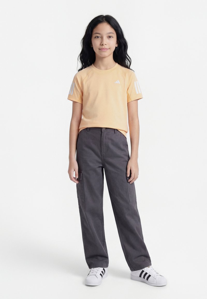 Κοπέλα με μακριά μαύρα μαλλιά στέκεται φορώντας ροδακινί κοντομάνικο μπλουζάκι, σκούρο γκρι παντελόνι cargo και λευκά αθλητικά παπούτσια με μαύρες ρίγες.