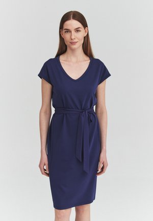 Femme aux cheveux bruns raides portant une robe bleu marine à col en V, manches courtes et ceinture nouée à la taille, debout devant un fond clair uni.