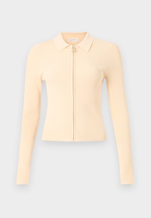 Frau mit blassgelbem langärmligem Zip-Up-Top, weißen weiten Hosen und schwarzen offenen High Heels, steht mit einer Hand in der Tasche.