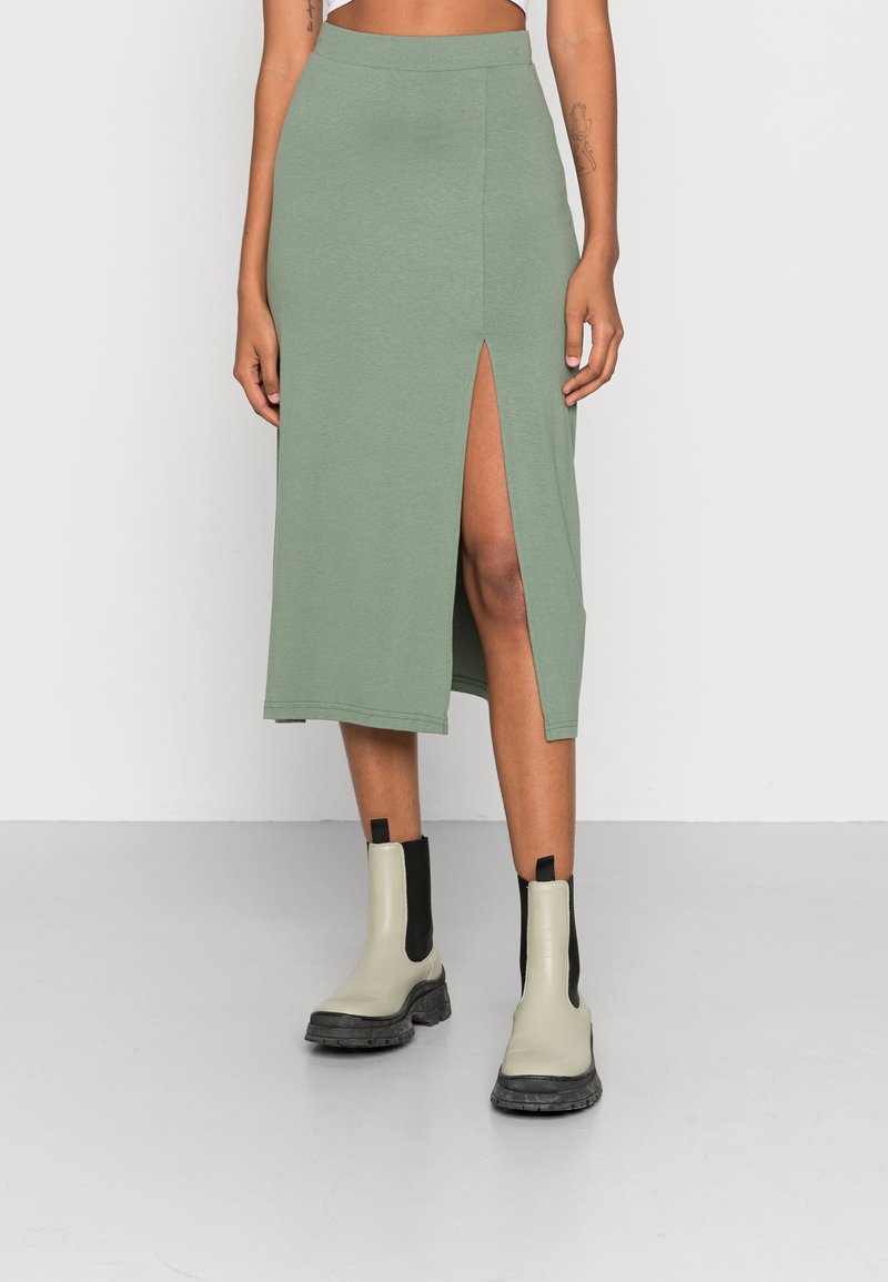 Mujer con falda midi verde con una abertura frontal y botas tobilleras gruesas de color gris claro, de pie contra un fondo blanco liso.