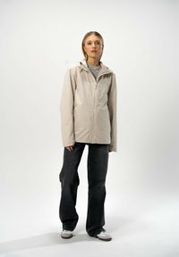 Beige vedenkestävä takki, jossa on huppu, vetoketjukiinnitys ja säädettävät nyörit. Yhdistetty leveälahkeisten tummien farkkujen ja valkoisten lenkkareiden kanssa.