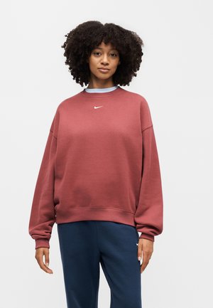Kvinde med krøllet hår iført en rustfarvet Nike sweatshirt og marineblå Nike bukser, stående mod en ensfarvet lys baggrund.
