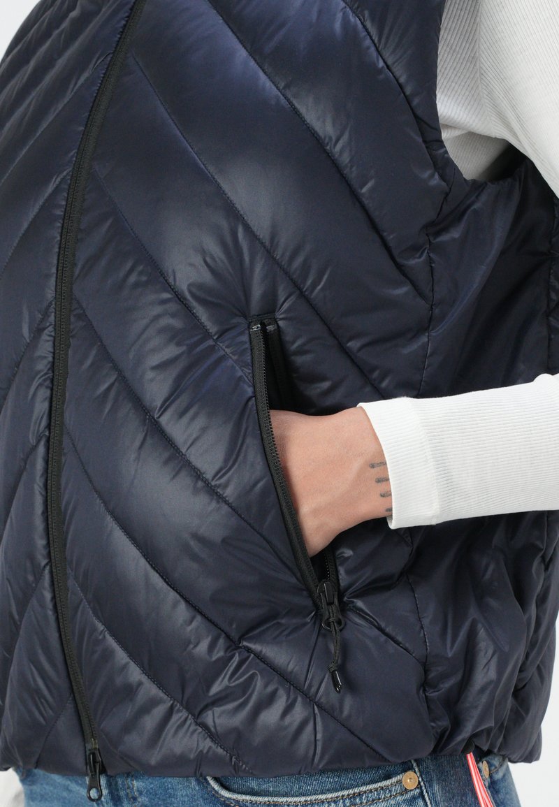 Marineblauer Puffer-Weste mit diagonaler Steppung, Reißverschlusstaschen und glatter Oberfläche. Darunter wird ein weißes Langarmshirt getragen.