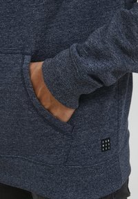 Sötétkék pulóver, textúrált anyagból, elülső zsebbel, bordázott mandzsettákkal és egy kis címkével, amelyen a "DNM WRKRS" felirat található az oldalon.