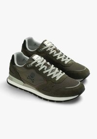 Olivgrüne Sneakers aus Wildleder und Mesh-Materialien. Merkmale eine kontrastierende weiße Zwischensohle, graue Schnürsenkel und ein Logo mit einem Totenkopf und gekreuzten Knochen.