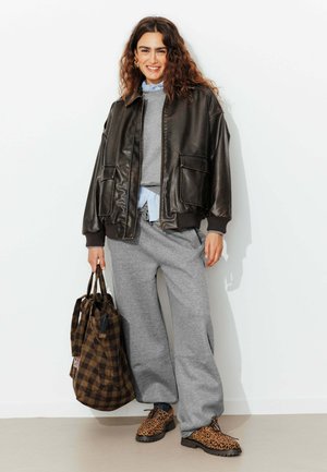 Veste bomber en cuir noir, sweatshirt gris, pantalon de survêtement gris, sac fourre-tout à carreaux marron, chaussures imprimé léopard, avec des cheveux ondulés, se tenant devant un fond blanc.