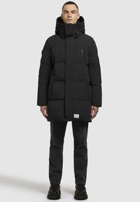 Mustmust järgnev rüü kõrge kraega, millel on lukuga taskud, ees nööpidega sulgemine ja ribakätised, kantud tumedate teksade ja saapatega.