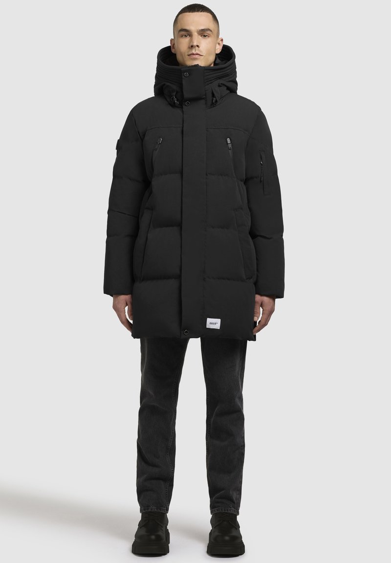 Sort puffersjakke med høj krave, der har lynlåslommer, knaplukning foran og ribbede mansjetter, båret over mørke jeans og støvler.