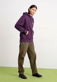 Sweat à capuche violet avec poche kangourou, associé à un pantalon vert et des chaussures noires. Le sweat à capuche présente un petit logo sur la poitrine.