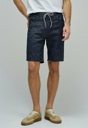 Hombre vestido con pantalones cortos de mezclilla oscuros con cintura de cordón, camiseta blanca y zapatillas beige, de pie contra un fondo liso.