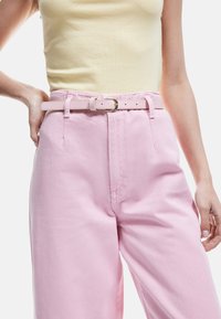 Pantalones rosa de tiro alto con una textura suave, que presentan un cinturón delgado a juego y una hebilla metálica. La parte superior es una camiseta sin mangas ajustada de color amarillo.