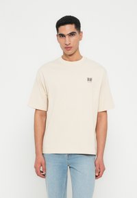 Junger Mann mit kurzen schwarzen Haaren, trägt ein beiges übergroßes T-Shirt mit kleiner schwarzer Schrift und hellblauen Jeans, steht vor einem weißen Hintergrund.