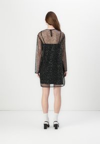 Rochie neagră, transparentă, cu mâneci lungi și o textură strălucitoare. Are o căptușeală ajustată, spate deschis și un închizător cu gât în formă de cheie.