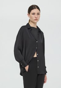 Camicia nera dal taglio morbido, maniche lunghe, chiusura frontale con bottoni e una texture liscia e lucida. Abbinata a pantaloni neri a vita alta.