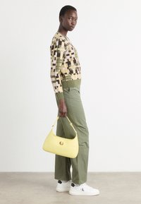 Pull en tricot avec un motif géométrique en beige, vert et marron ; pantalon vert olive ; sac à main jaune ; baskets blanches avec des accents noirs.