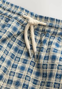Tissu à carreaux bleu et blanc avec une ceinture élastique froncée et un cordon épais de couleur crème noué en un nœud avec des embouts en métal.