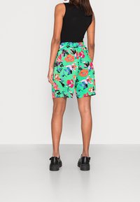 Shorts fleuris en vert vif, ornés de grandes fleurs roses et orange. Tissu léger avec une taille élastique et une coupe décontractée. Chaussures noires.