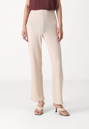 Pantalons côtelés beige clair avec taille haute et jambes larges. Le tissu a une texture douce. Associés à des sandales légères.