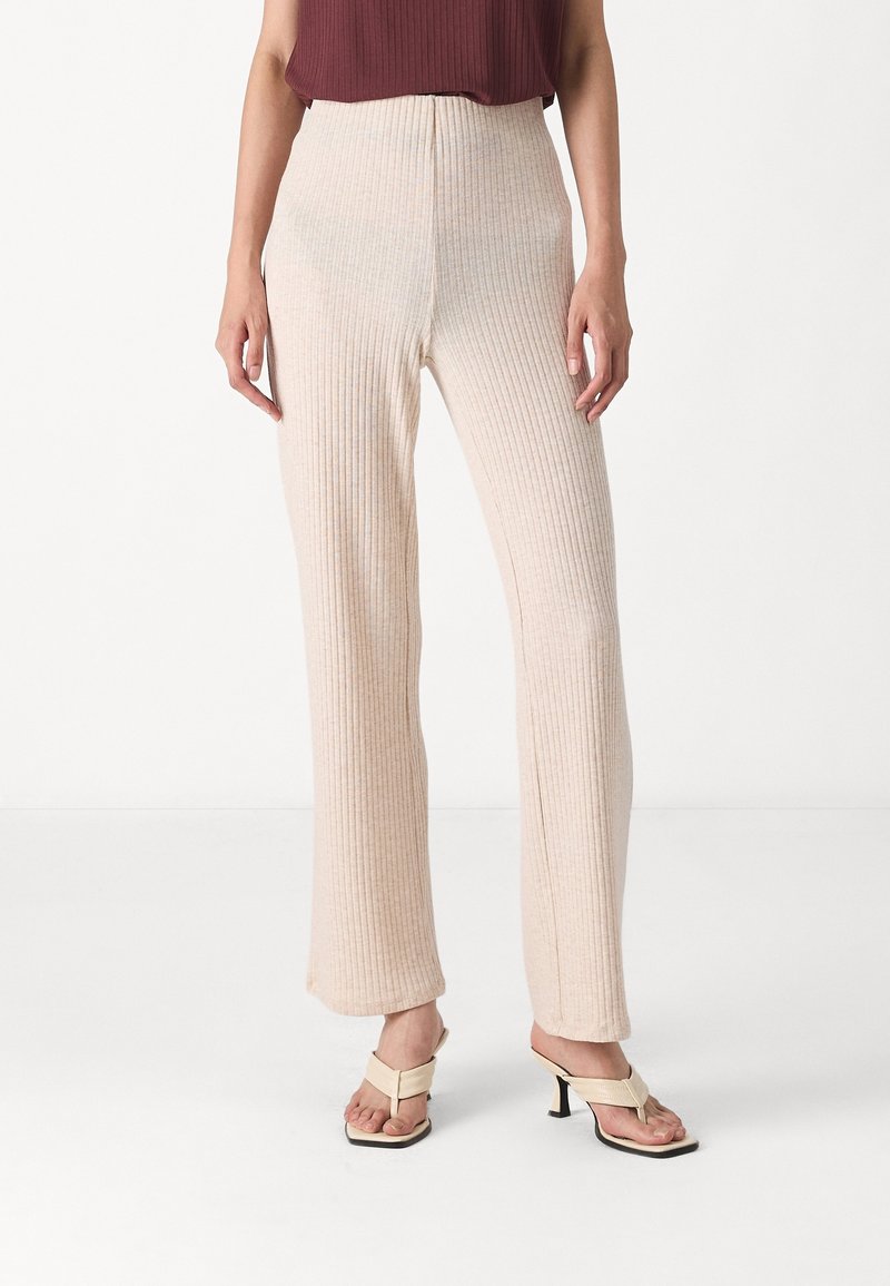 Ljust beige ribbat stickade byxor med hög midja och vida ben. Tyget har en mjuk textur. Kombinerat med ljusa sandaler.