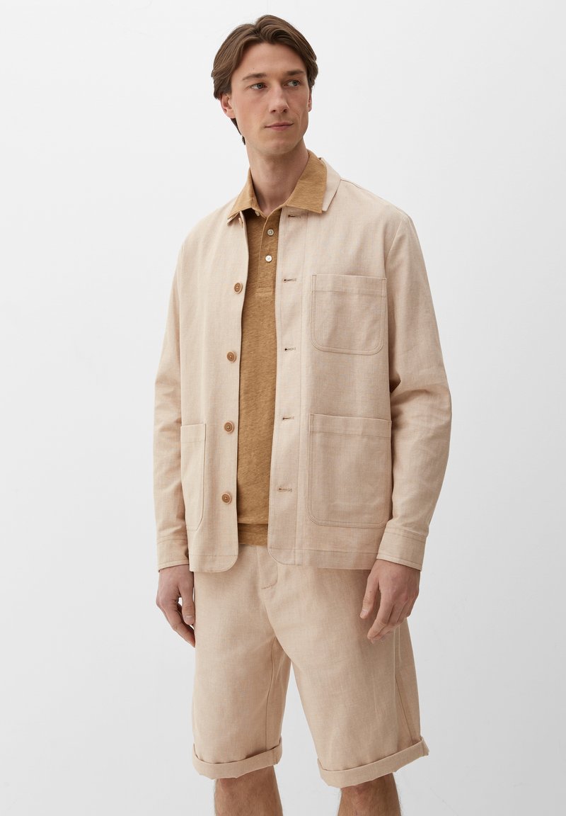 Jasnobeżowa kurtka o kołnierzowym fasonie, z dwoma przednimi kieszeniami, zapięciem na guziki i miękką teksturą, zestawiona z brązowym polo i szortami.