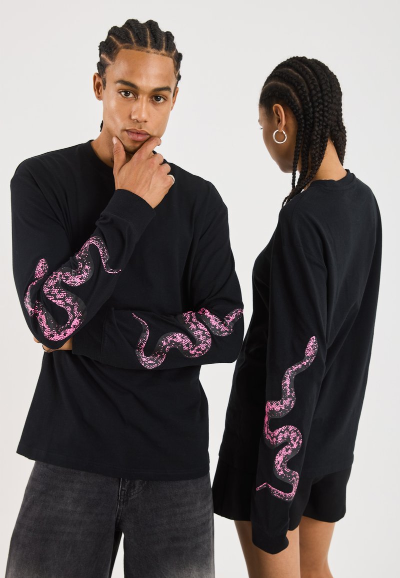 Chemise noire à manches longues avec un graphique de serpent rose le long des manches. Fabriquée en coton, avec une coupe décontractée et un col rond.
