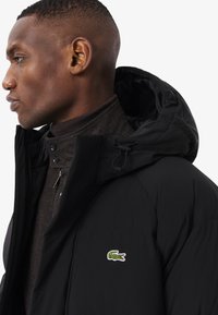 Chaqueta acolchada negra con capucha, que presenta un logo de cocodrilo Lacoste en verde y blanco en el pecho izquierdo y un cuello alto.