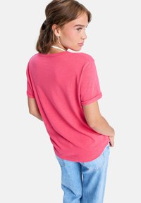 Ung kvinde med brunt hår iført en lyserød kortærmet skjorte og lyseblå jeans, der kigger over skulderen mod en hvid baggrund.