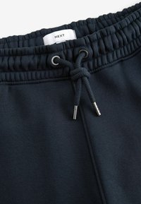 Los pantalones de chándal azul marino presentan una cinturilla elástica con cordón, aglets metálicos y una superficie texturizada de tela. Etiqueta de la marca visible.