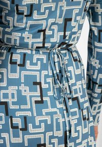 Geometrinen kuvioinen mekko sinisestä kankaasta; siinä on valkoisia ja mustia muotoja, vyötärö on kireä ja siinä on yhteensopiva solki. Sujuva pinta ja kevyt muotoilu.