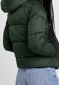 Person som bär en mörkgrön puffjacka med huva och ljusblå högmidjade jeans, stående mot en enkel bakgrund.