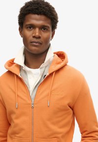 Orange zip-up hoodie lavet af bomuld med et gråt indre lag, der har et logo på brystet og snøre i hætten.
