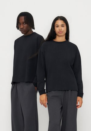 Deux jeunes adultes portant des hauts noirs à manches longues et des pantalons gris foncé se tiennent côte à côte devant un fond clair uni.