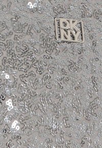 Srebrna tkanina z cekinami o teksturowej powierzchni i falistym wzorze. W dolnym prawym rogu znajduje się małe logo DKNY.