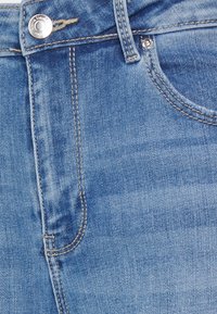 Jeans de denim azul com um bolso frontal, costura visível e um detalhe em botão metálico na cintura. Textura suave com um acabamento desbotado.