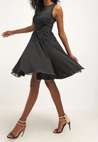 Svart klänning med spetsliv, utsvängd chiffongkjol och ärmlös design. Sandaler med strappy i nude kompletterar outfiten.