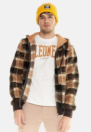 Jeune homme portant un bonnet jaune, un t-shirt blanc "LEONE 1947" et une veste zippée à carreaux marron avec un pantalon beige, debout sur fond blanc.