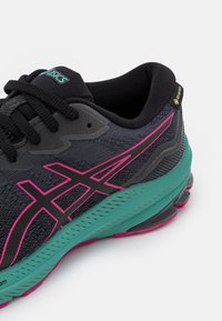 Chaussure de sport noire avec une tige texturée, des accents roses et turquoises, et une semelle rembourrée au design incurvé distinctif.