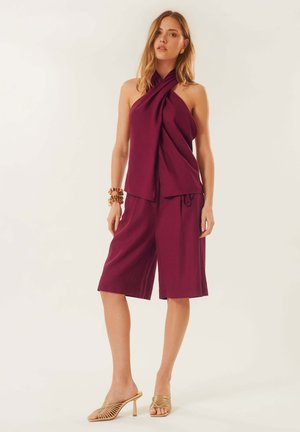Femme portant un haut sans manches prune à col halter et un short assorti longueur genou, accessoirisée de bracelets dorés et de sandales dorées à talons.