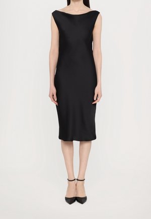 Vrouw draagt een mouwloze, knielange zwarte jurk met een boothals en zwarte puntige enkelsandalen met enkelriempje, staand tegen een witte achtergrond.